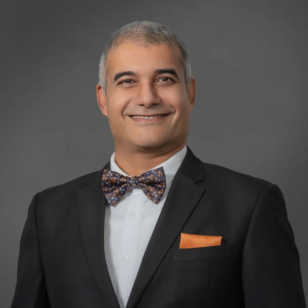 Person in a black suit jacket and white dress shirt, wearing a dark floral bow tie and an orange pocket square against a gray studio backdrop.