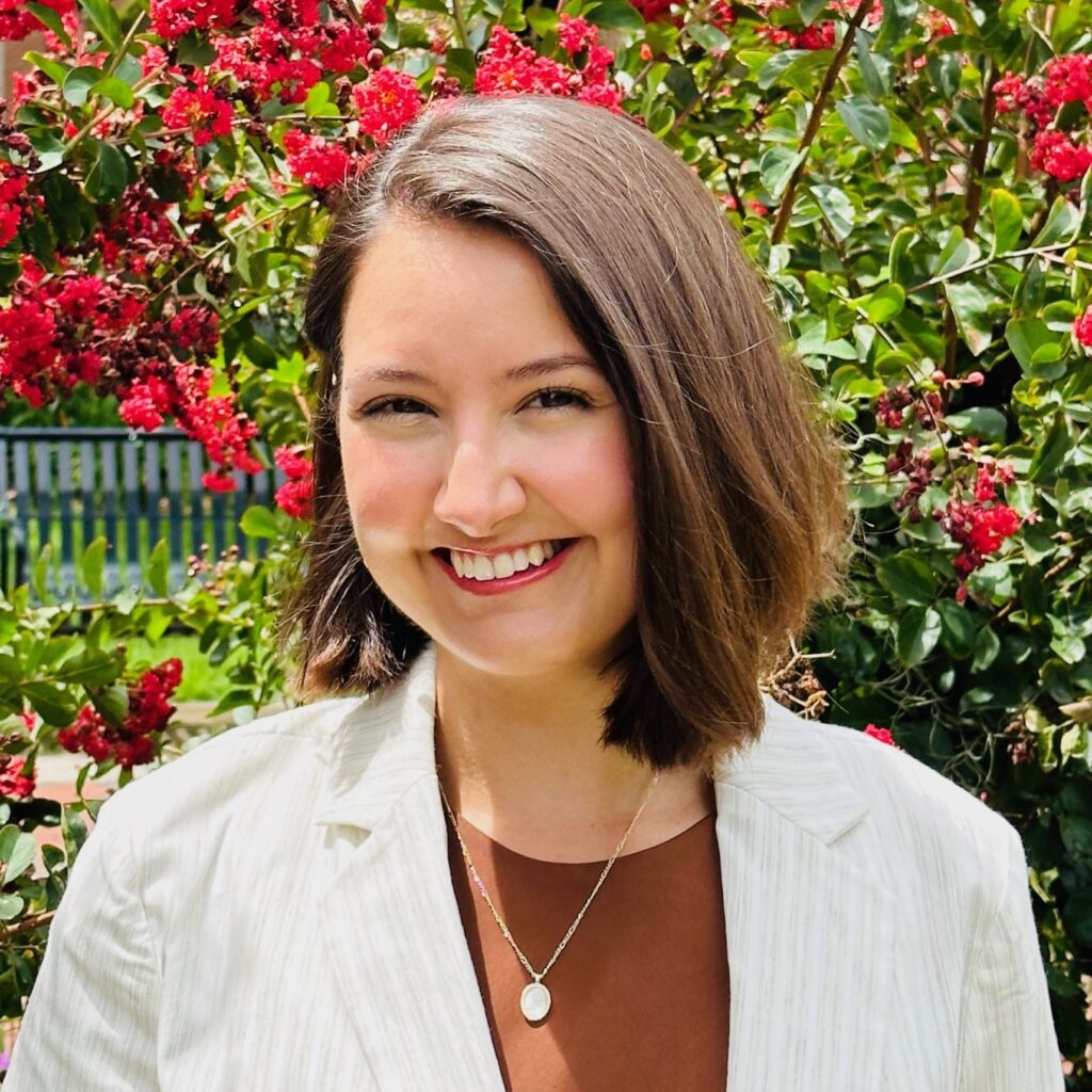 Person with shoulder-length brown hair wearing a light pinstriped blazer, brown top, and oval pendant necklace, standing in a sunny garden with bright red flowers and green foliage, with a bench in the background.