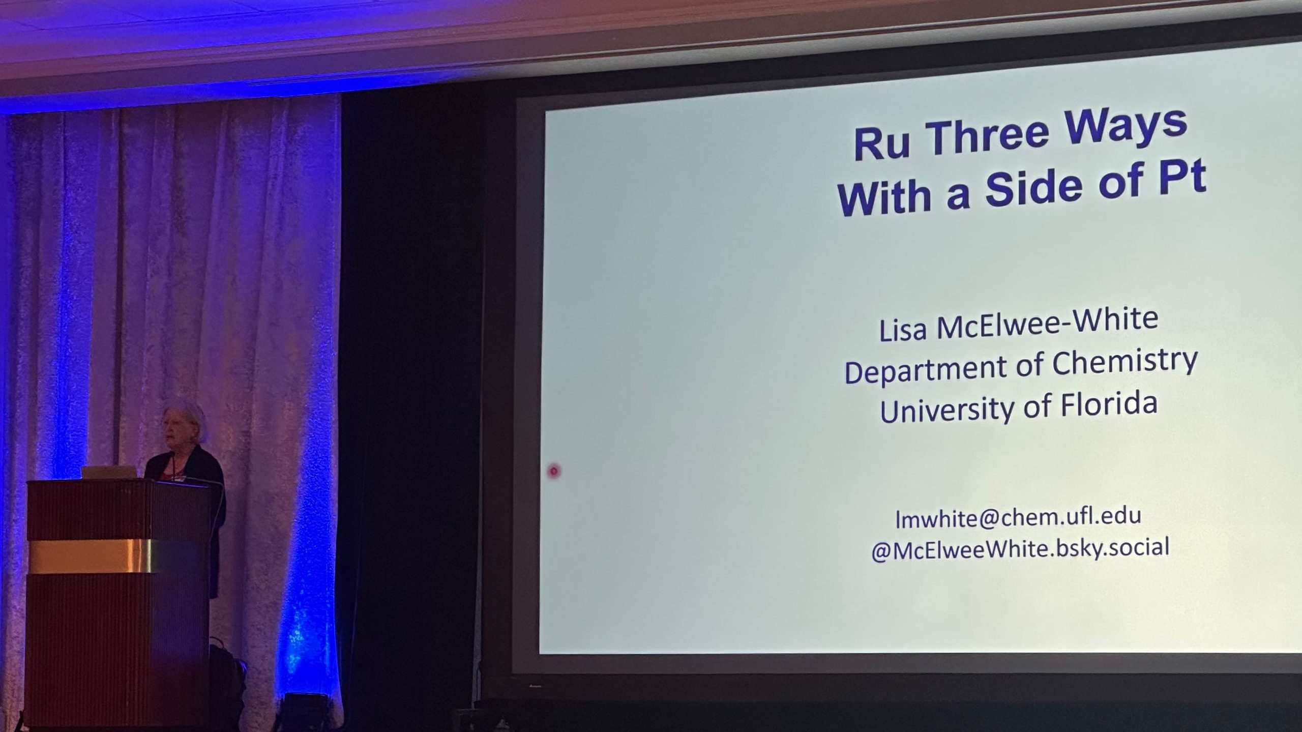 Prof. Lisa McElwee-White stands at a podium beside a large screen displaying the title of her plenary lecture, “Ru Three Ways with a Side of Pt,” at FAME 2026.